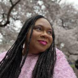 A headshot of an artist wearing a pink sweater surrounded by blossoming trees